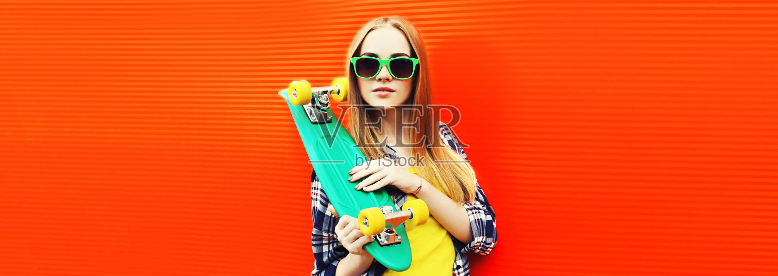 现代年轻的金发女郎肖像与绿色滑板戴墨镜在彩色的红色背景照片摄影图片