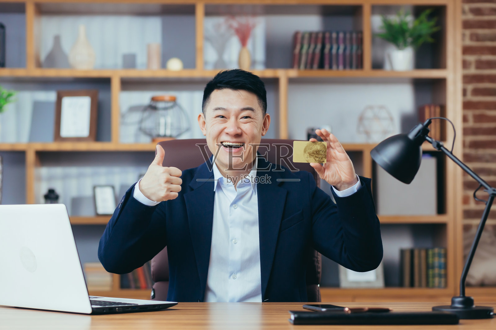 一个成功的亚洲商人的肖像在一个经典的办公室，看着相机，微笑着拿着手机和银行信用卡，网上购物照片摄影图片