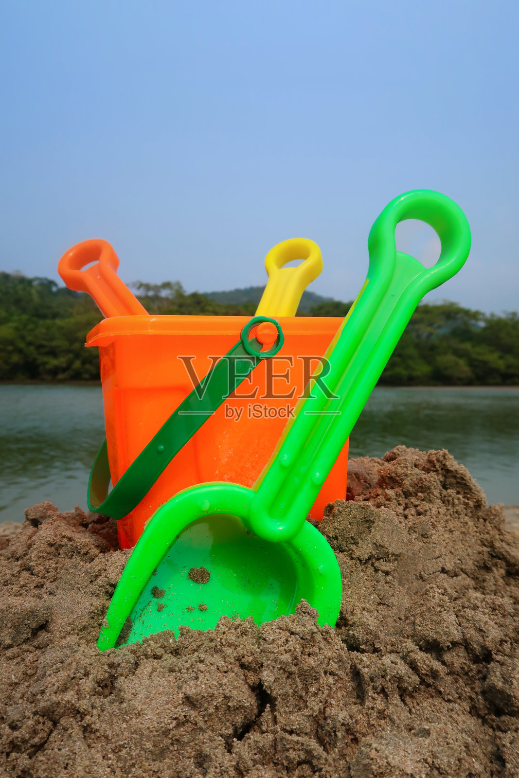 塑料桶，铲子和耙子在沙子堆上的特写图像，沙滩上鲜艳的儿童海滩玩具，大海和海岸线，林地与棕榈树，重点在前景照片摄影图片