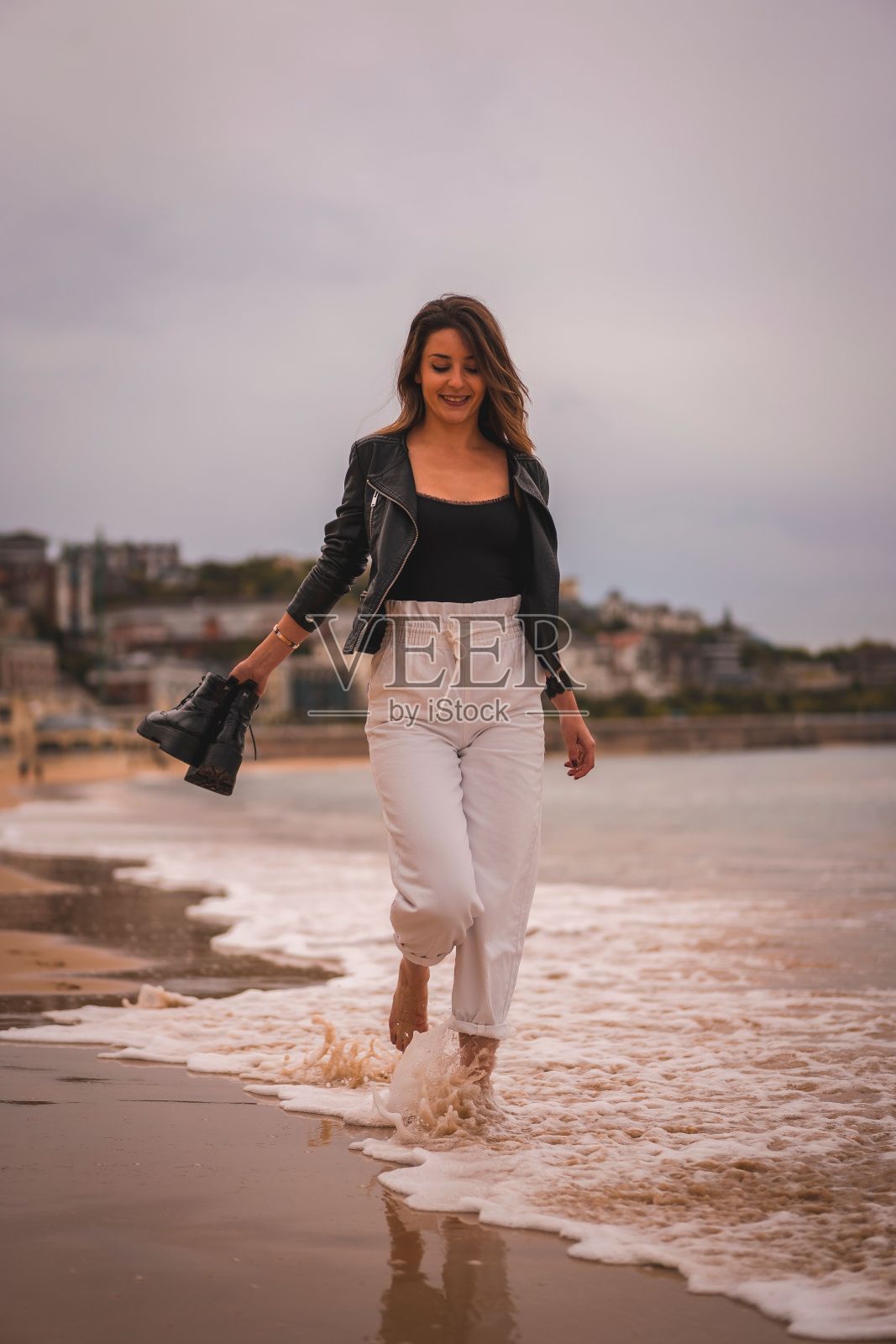 垂直拍摄的年轻时尚的女性在日落时赤脚走在海滩上，抱着她的海岸照片摄影图片