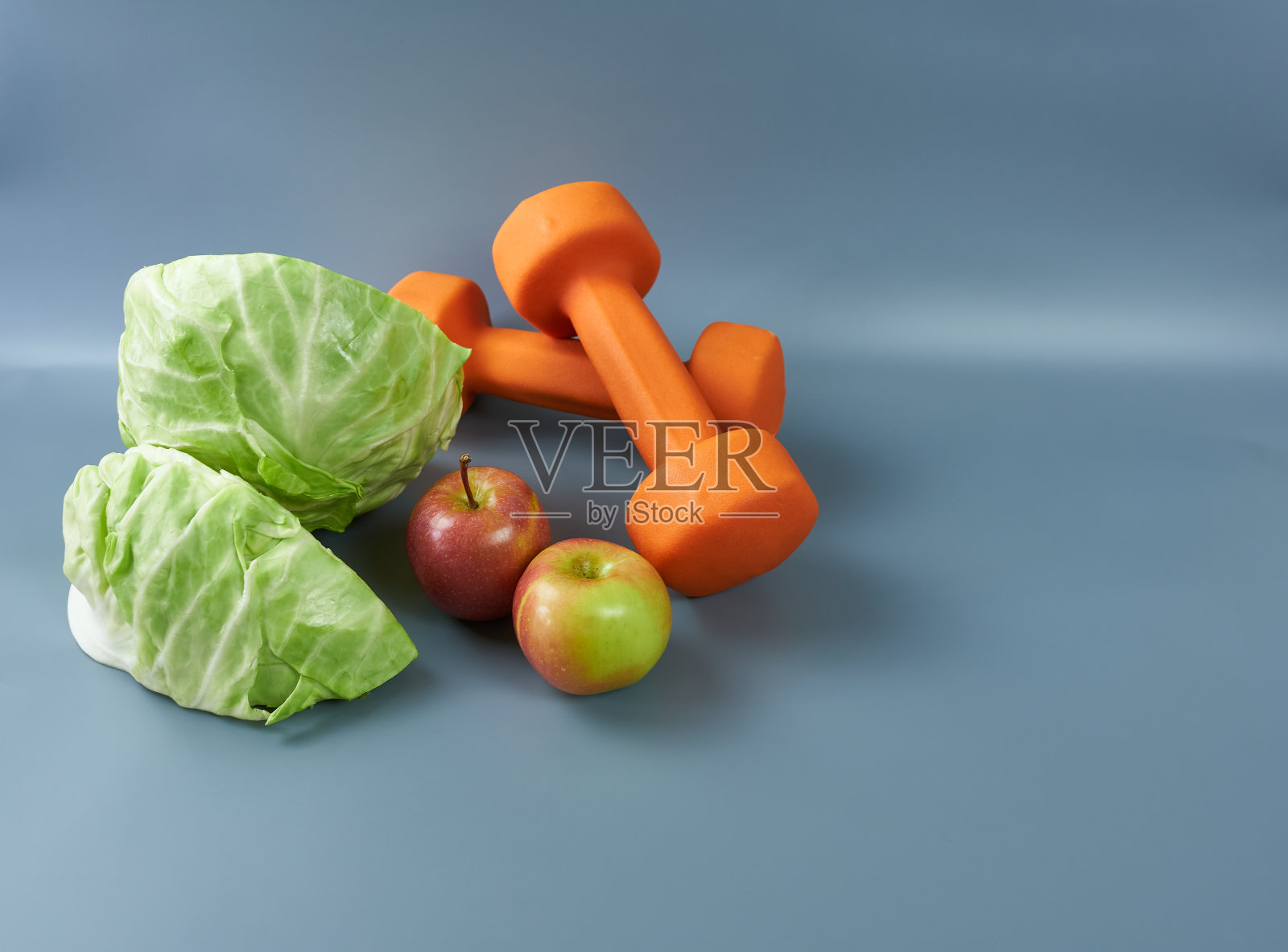 卷心菜、苹果和橙色的哑铃，在灰色背景上锻炼。这个概念就是锻炼和饮食。照片摄影图片