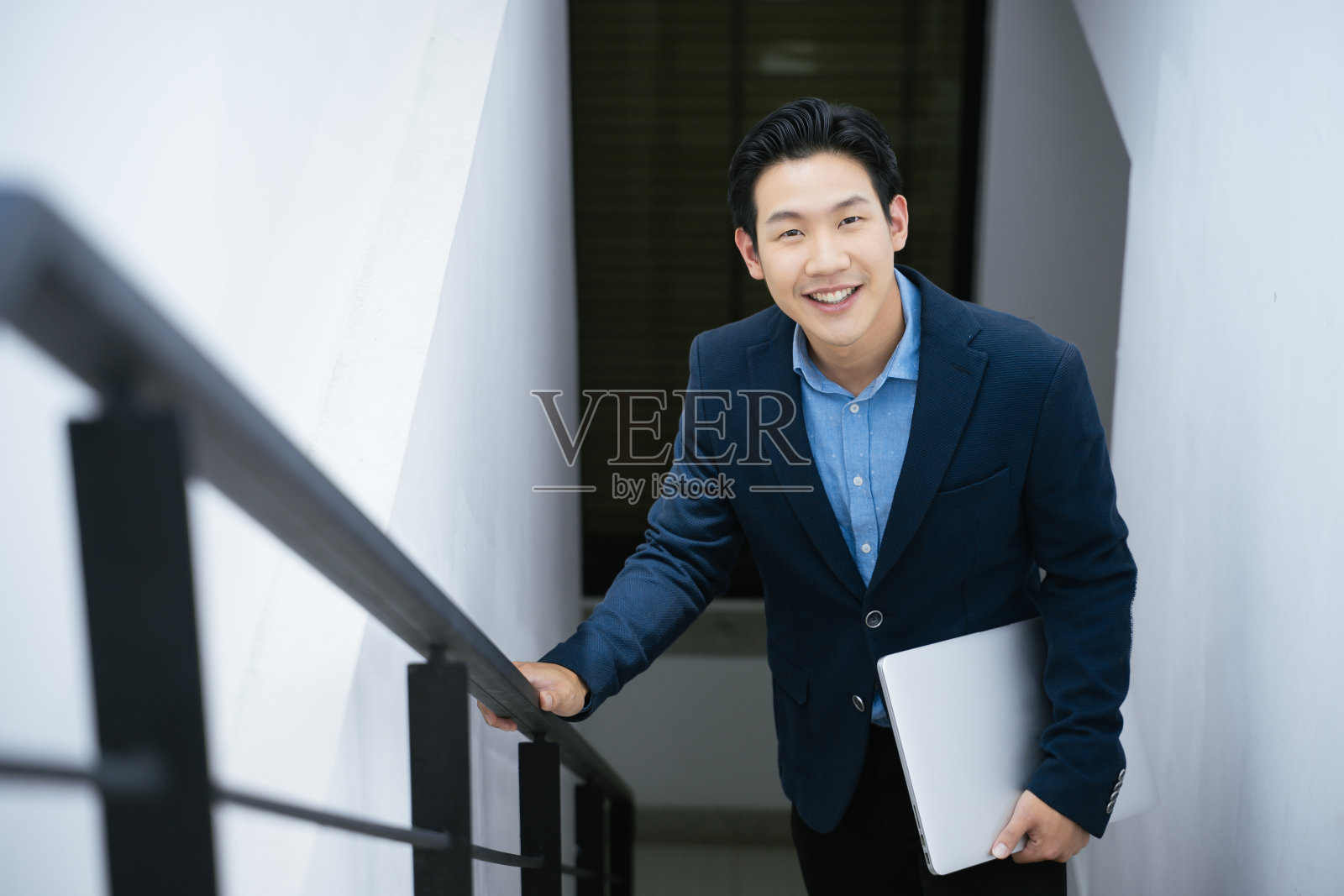 亚洲商人在办公室楼梯上自信地微笑，手里拿着笔记本电脑。照片摄影图片