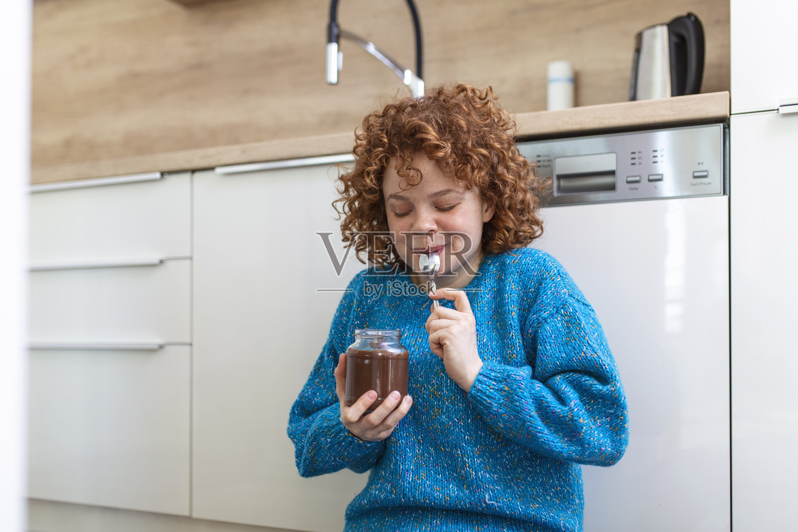 年轻女子坐在厨房地板上吃着罐子里的巧克力。照片摄影图片