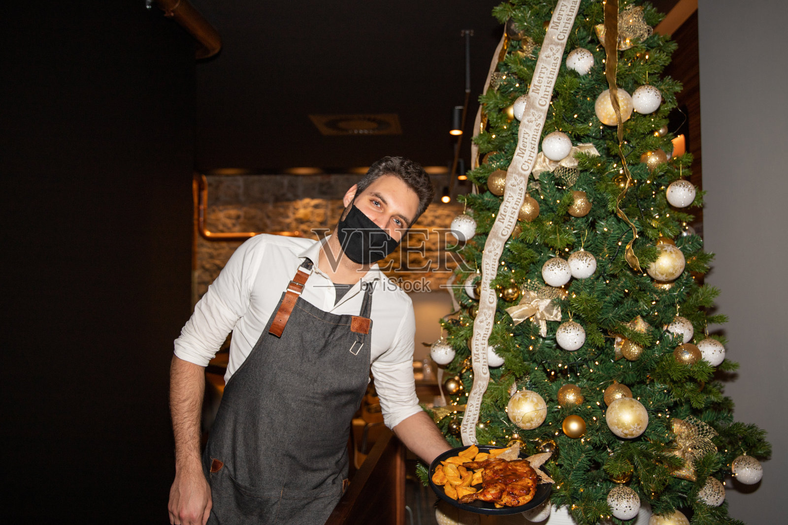 圣诞节期间，服务员端着盘子来到顾客的餐桌上，用安全面罩捂住口鼻照片摄影图片