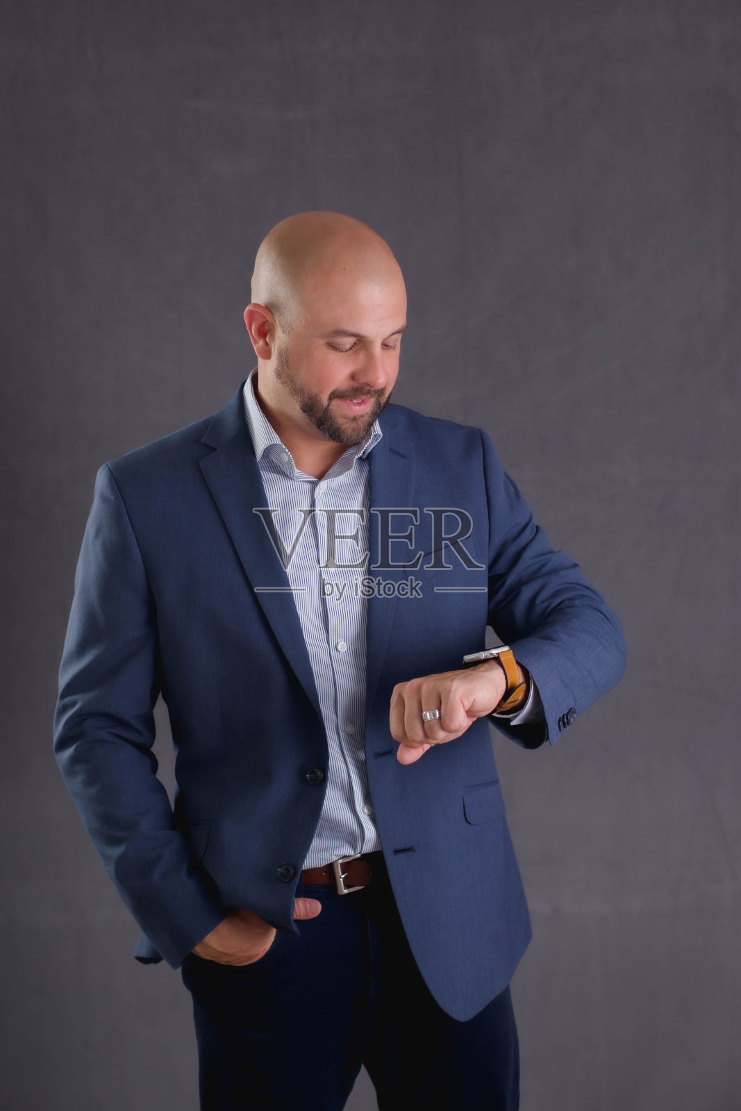 工作室拍摄的一个年轻的秃顶商人在西装检查时间站在灰色背景照片摄影图片