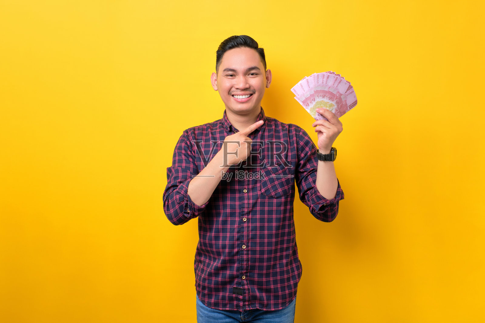 微笑的年轻亚洲男子指着钞票，赚取现金在网上孤立的黄色背景。利润与财富概念照片摄影图片