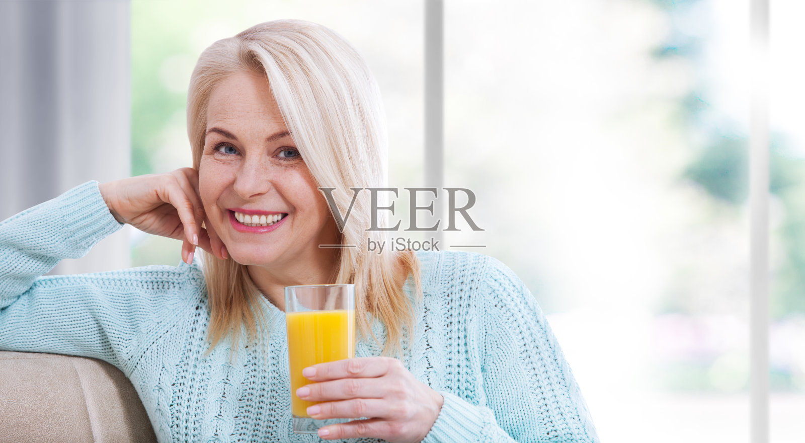 快乐的女人端着一杯橙汁，坐在窗边，对着镜头看起来很放松。照片摄影图片