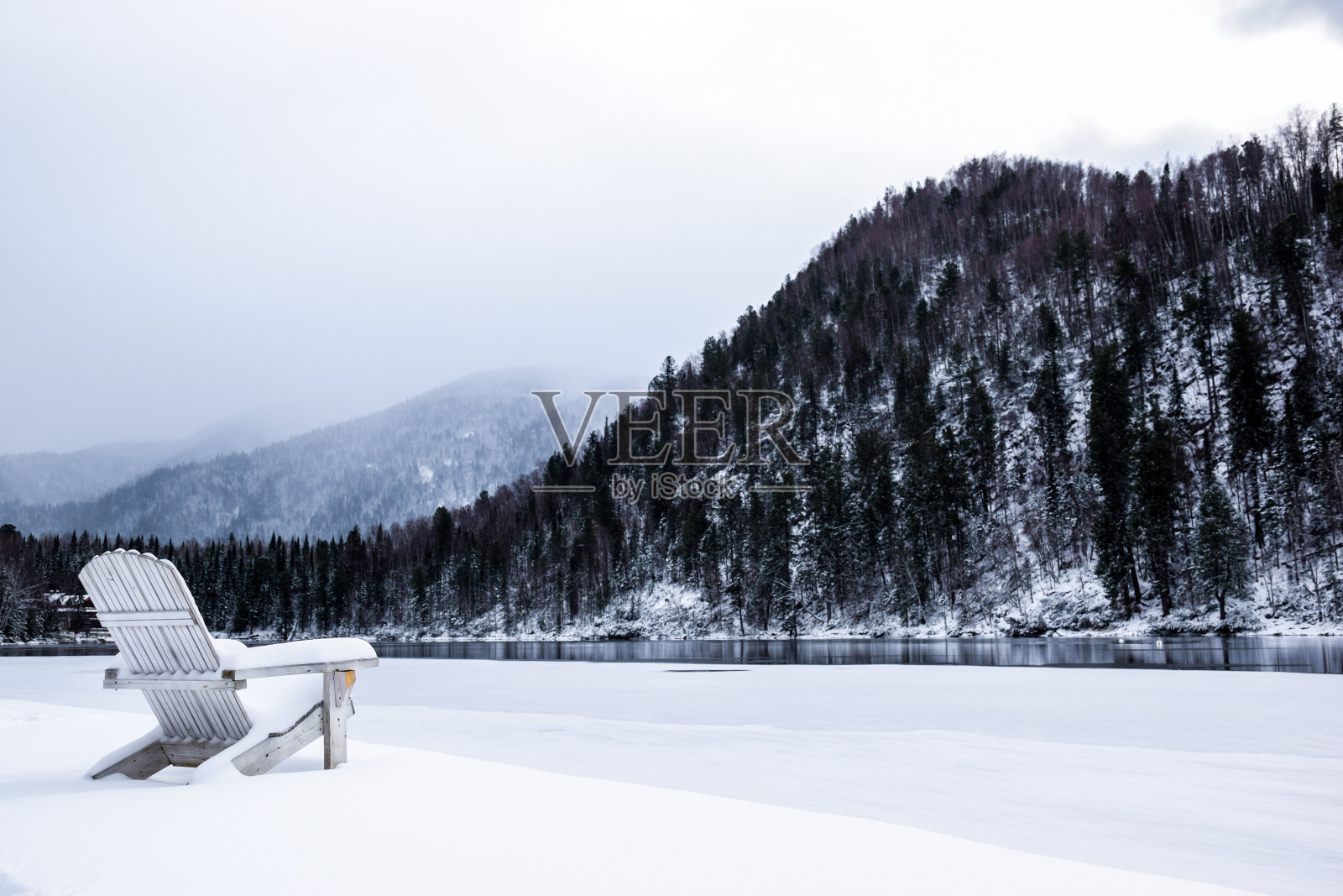 冬天湖边的度假胜地。冰湖边雪地里的木椅。湖岸边的雪地上有一把木椅，背景是群山。冬天湖边的雪椅。照片摄影图片