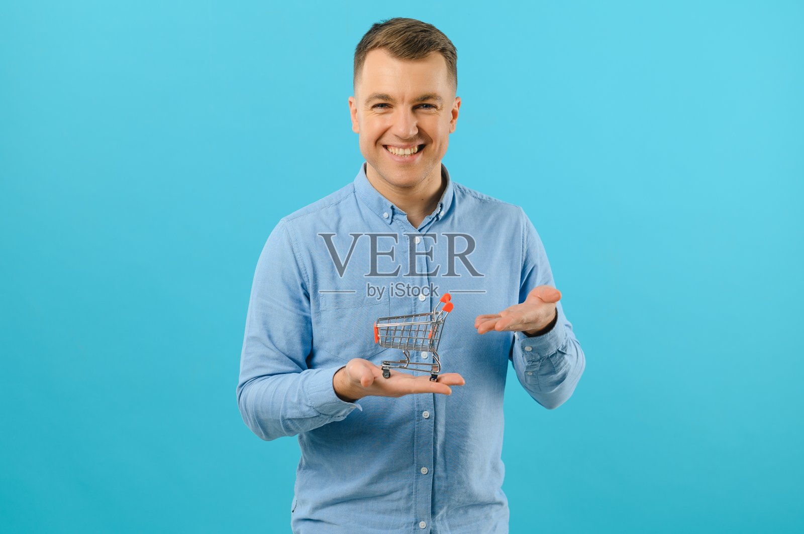 微笑的男子手持迷你购物篮在蓝色背景的肖像。增加销售的概念。照片摄影图片