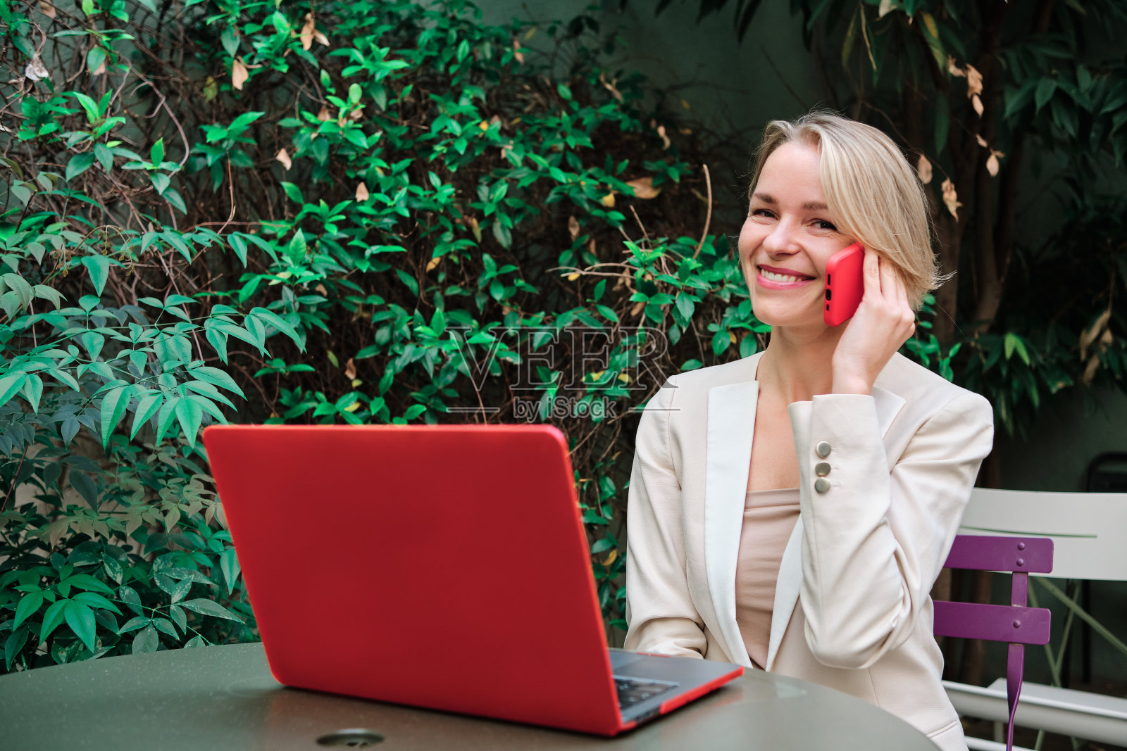 女商人坐在户外的桌子上，一边看着镜头，一边微笑着打电话。照片摄影图片