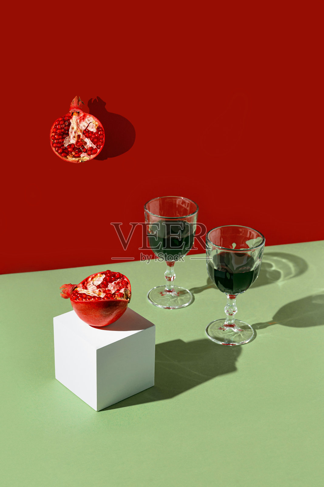由半石榴和酒杯在明亮的红色和绿色背景与阳光和阴影的创造性组成。夏日茶点概念照片摄影图片