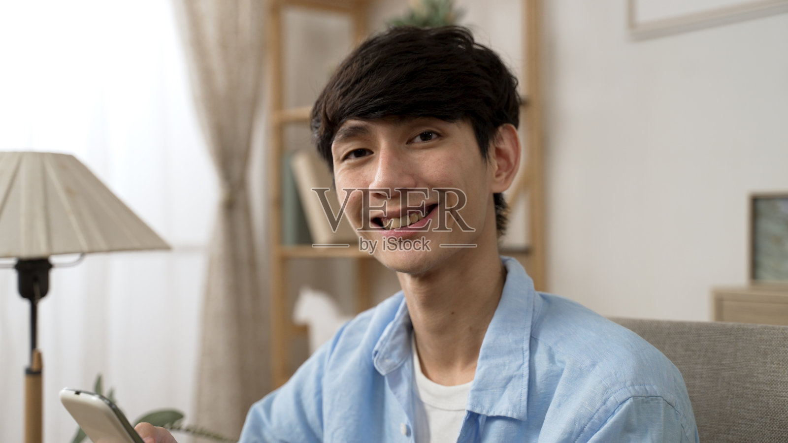 白天，在家里的客厅里，一名欢快的亚洲男子一边使用智能手机，一边转身微笑着看着相机。照片摄影图片