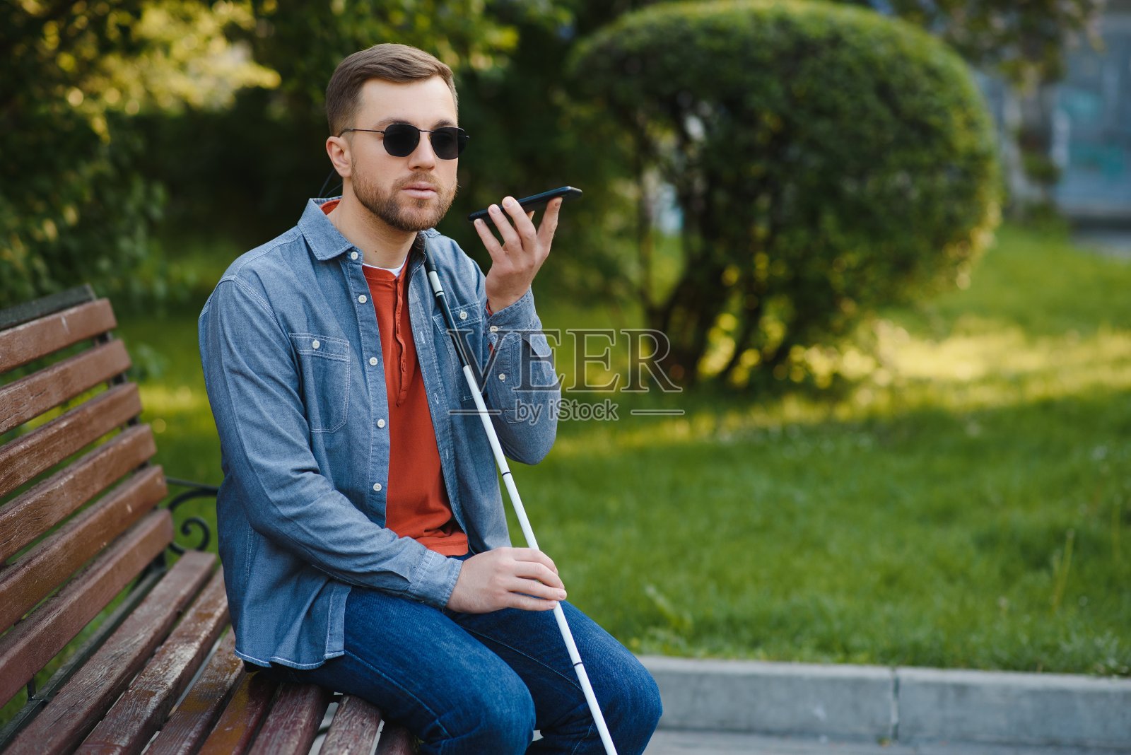 年轻的盲人拿着智能手机坐在城市公园的长椅上打电话。照片摄影图片