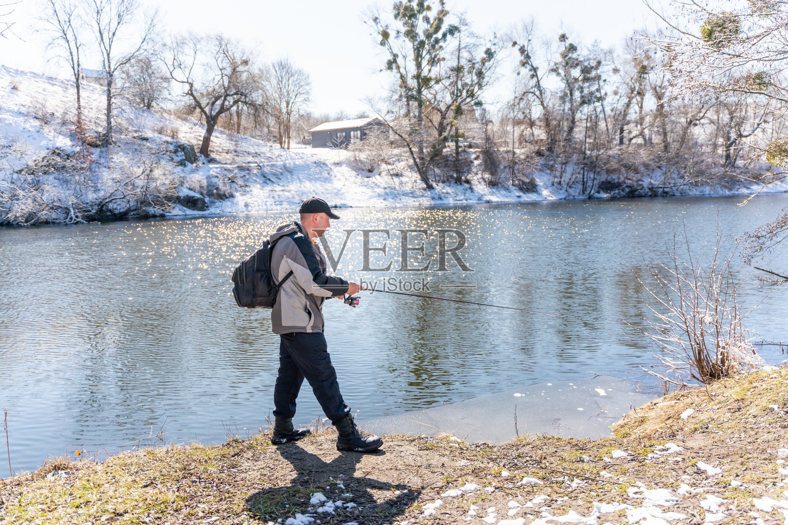 初春，一个渔夫背着背包，拿着鱼竿在白雪覆盖的河边捕鱼照片摄影图片