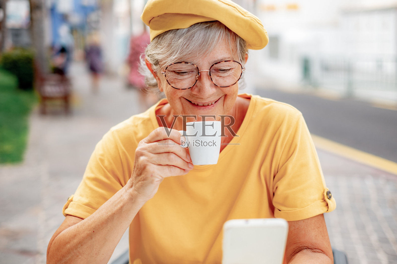 微笑的女人在黄色拿着咖啡杯坐在自助餐厅的桌子-老太太享受休息在早上照片摄影图片
