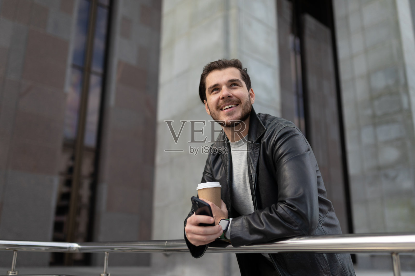 一个成年男子面带微笑，端着一杯咖啡，拿着手机，抬起头来照片摄影图片