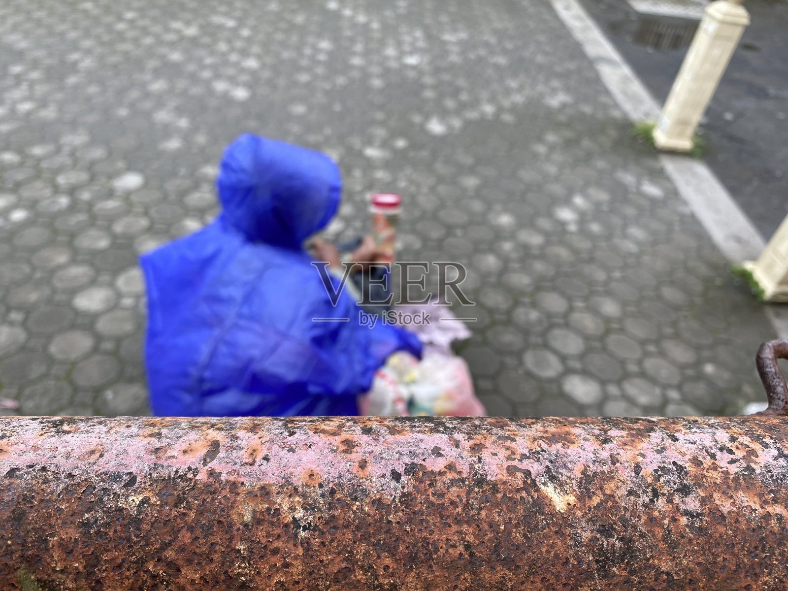 穿着蓝色雨衣的乞丐坐在教堂墓地的人行道上挥舞着施舍的玻璃杯。照片摄影图片