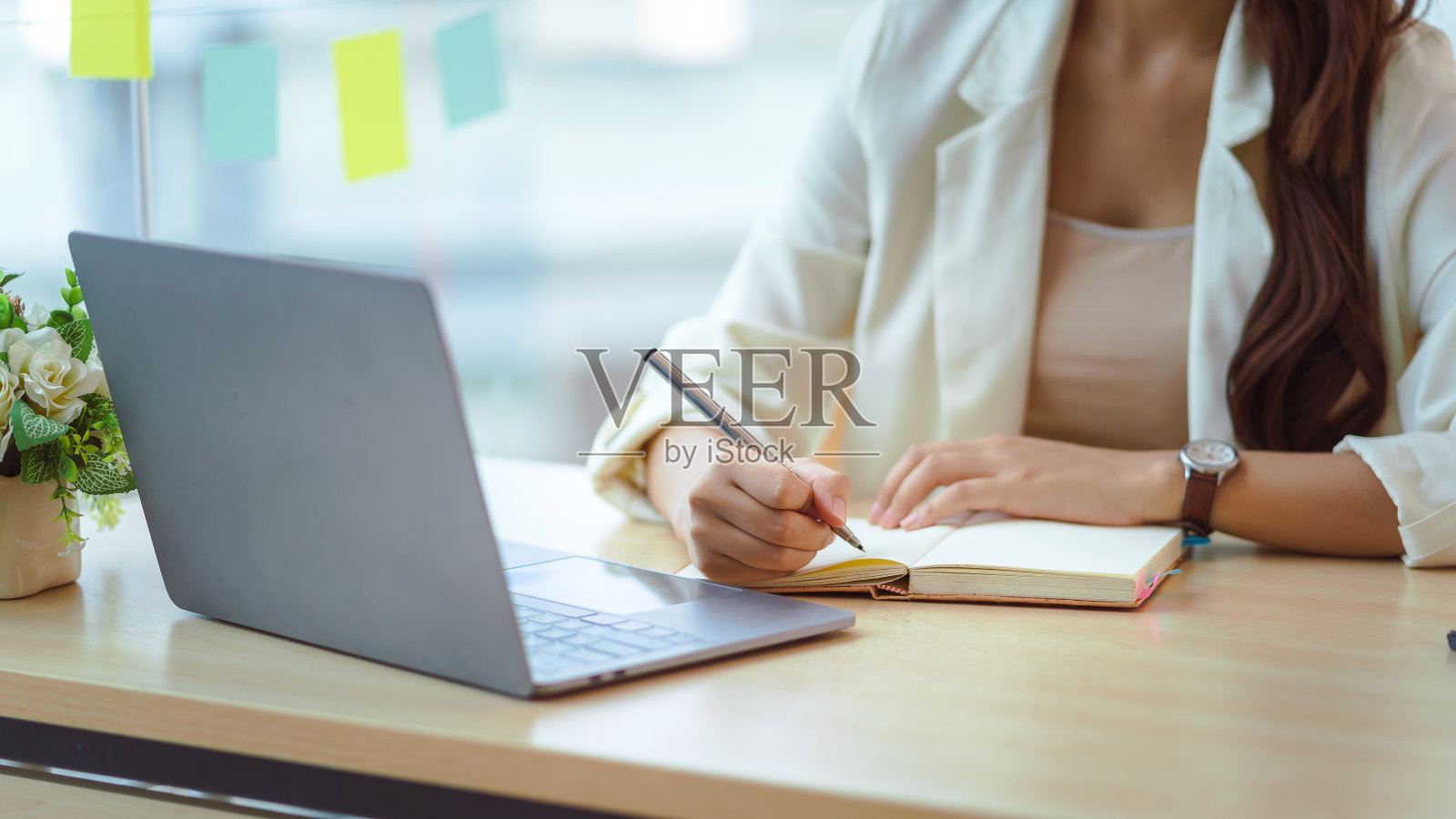 一个女人在办公室用笔记本电脑在笔记本上写字和记笔记的特写图像。照片摄影图片