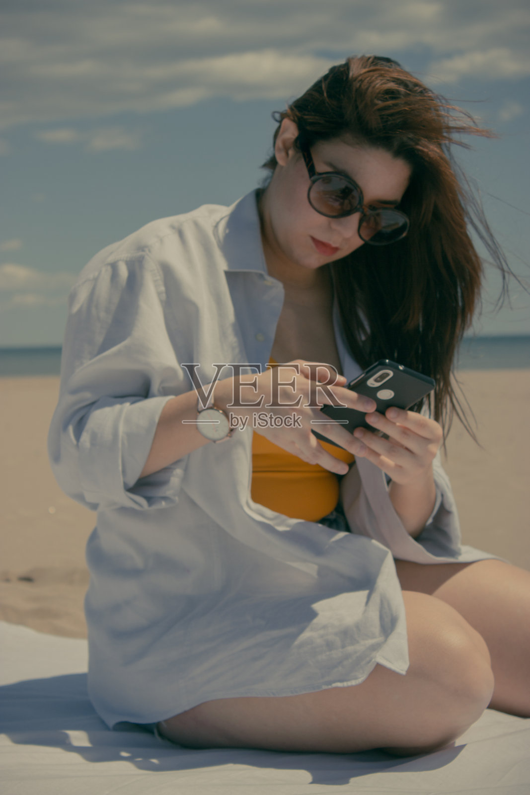 浅聚焦一个年轻的棕发西班牙女子使用她的手机在海滩上被大海包围照片摄影图片