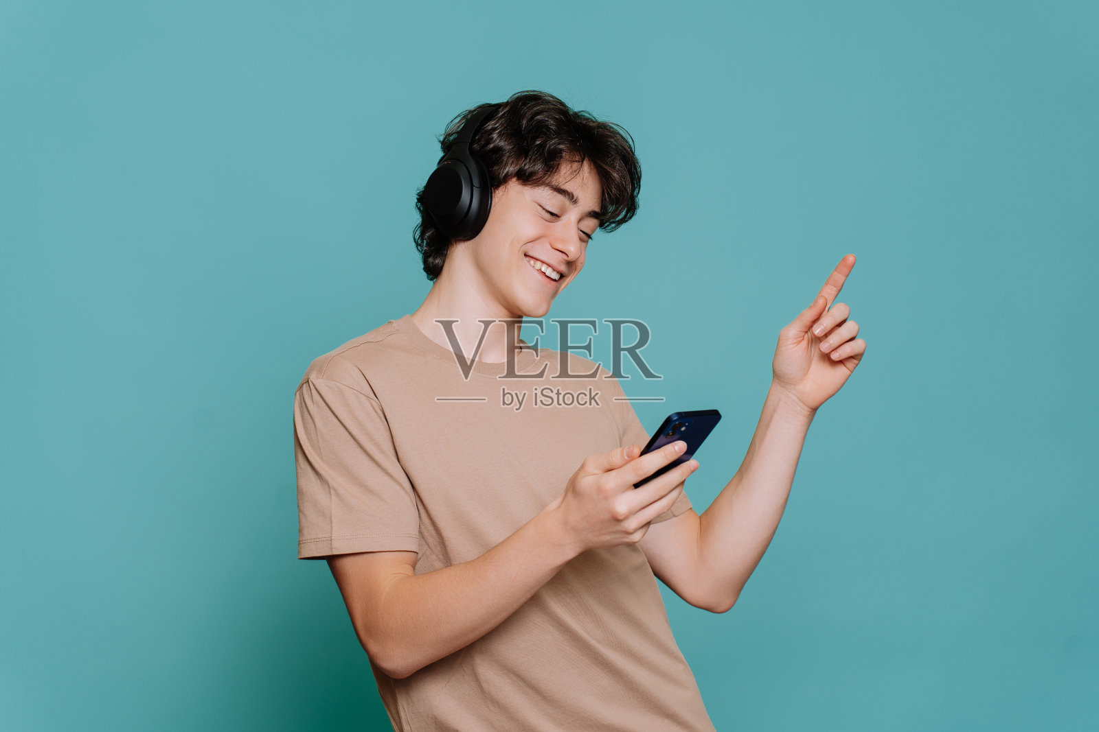 英俊的西班牙年轻人听音乐，用耳机握住手机，眼睛闭在蓝色的工作室背景。快乐的男孩跳舞，笑容灿烂。快乐的少年热爱音乐。模型、青年。照片摄影图片