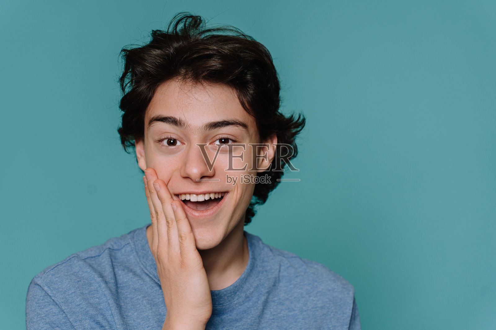 近距离的英俊的男孩棕色头发看着相机咧嘴笑，张开嘴触摸脸惊讶。白种人孤立在蓝绿色工作室背景。模型，坦率的情感。照片摄影图片