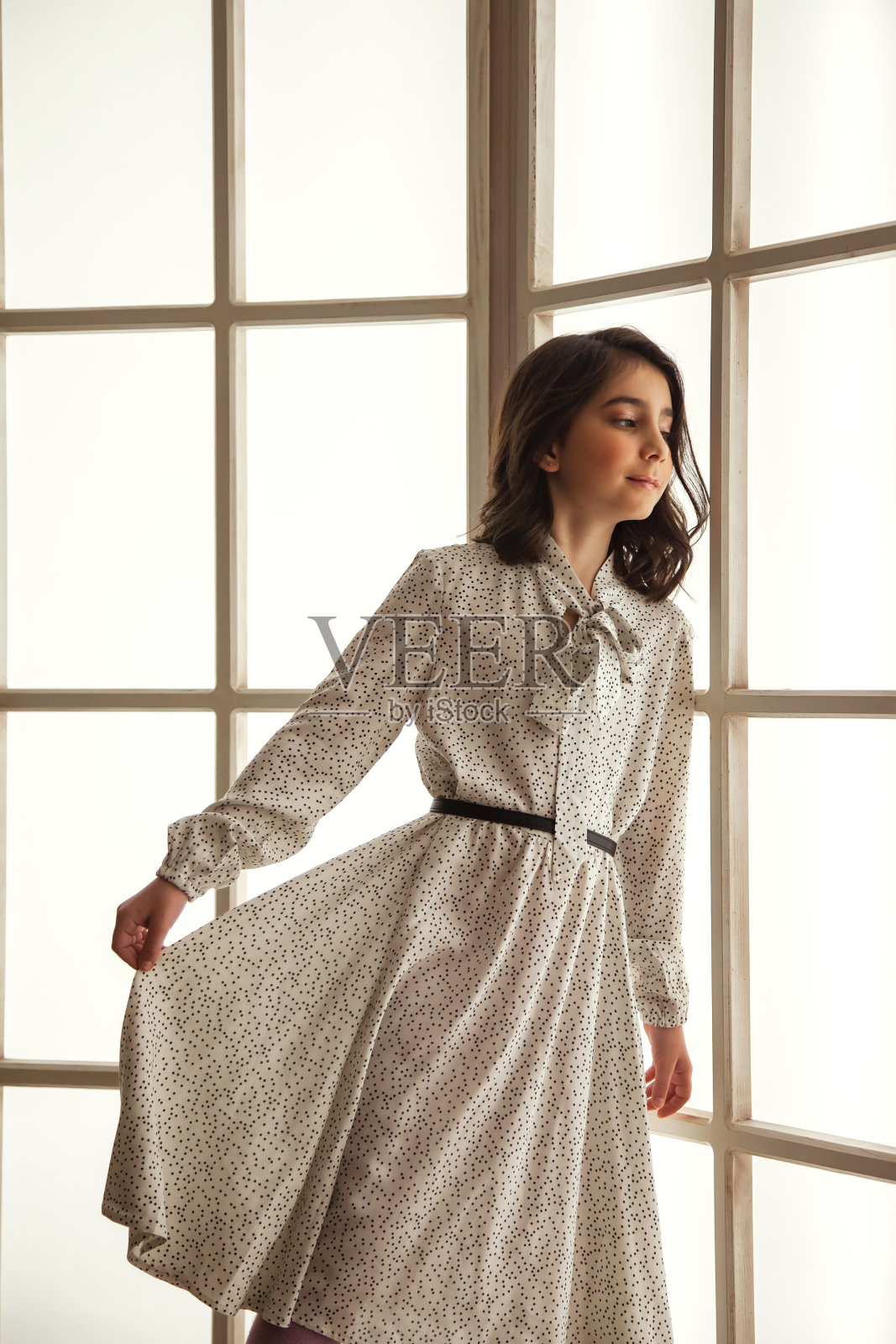 小时髦的女孩在白色连衣裙摆造型大光窗室内，看向别处。工作室拍摄了积极的青少年漂亮女士。儿童积极情绪概念。复制广告的文字空间照片摄影图片