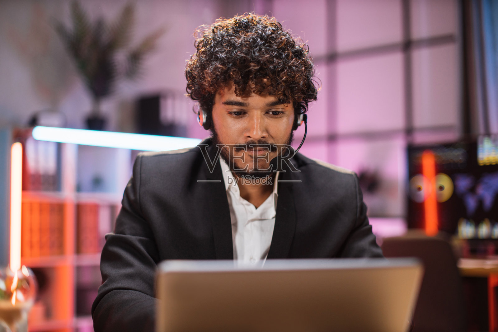 肖像自信的胡须男子在正式穿着和耳机工作在办公室使用笔记本电脑。照片摄影图片