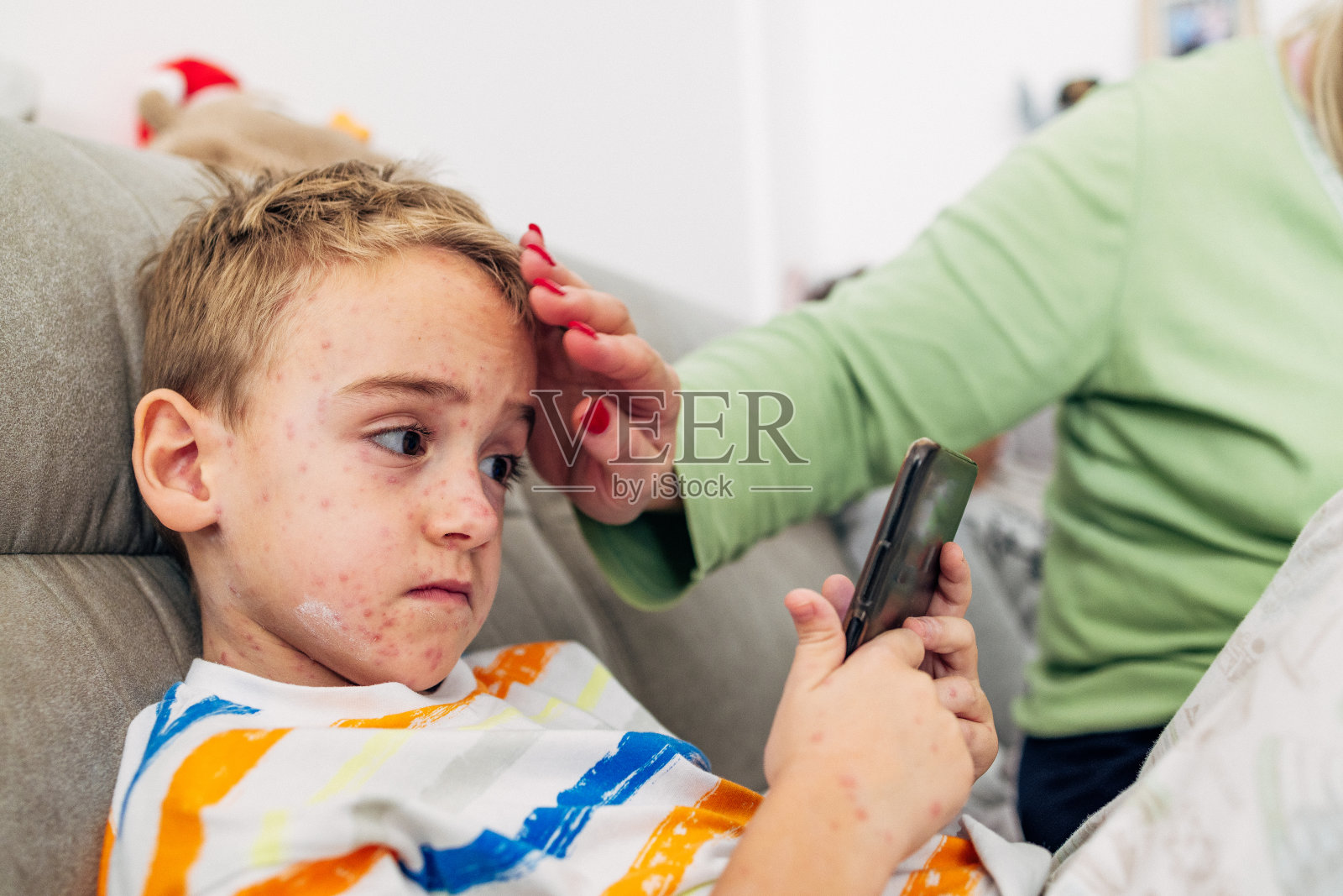 儿子玩手机游戏的时候，妈妈在他的皮肤上涂了一种乳液，以减少水痘的瘙痒照片摄影图片