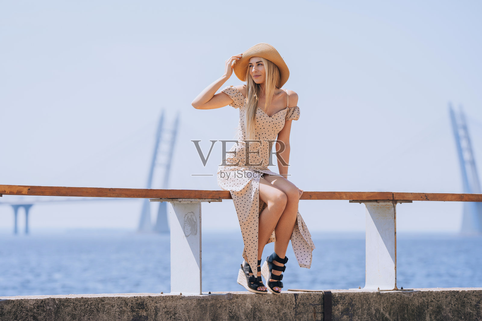 忧郁的年轻金发意大利女子坐在堤岸外穿着米色连衣裙，草帽看着远处的海边和桥的背景。漂亮的女士在旅行。夏季休闲。照片摄影图片