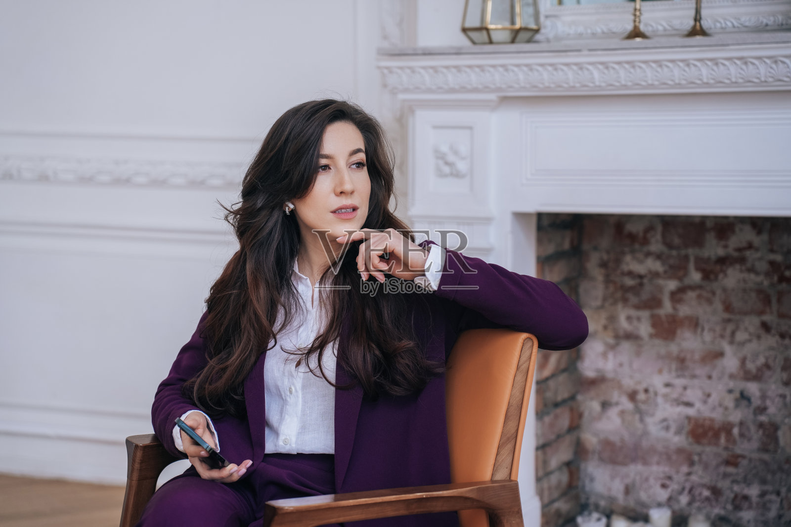 披着波浪卷发，身穿勃艮第西装，坐在椅子上，拿着手机，看向镜头的迷人商务女性肖像。疲惫的女企业家在办公室里沉思。照片摄影图片