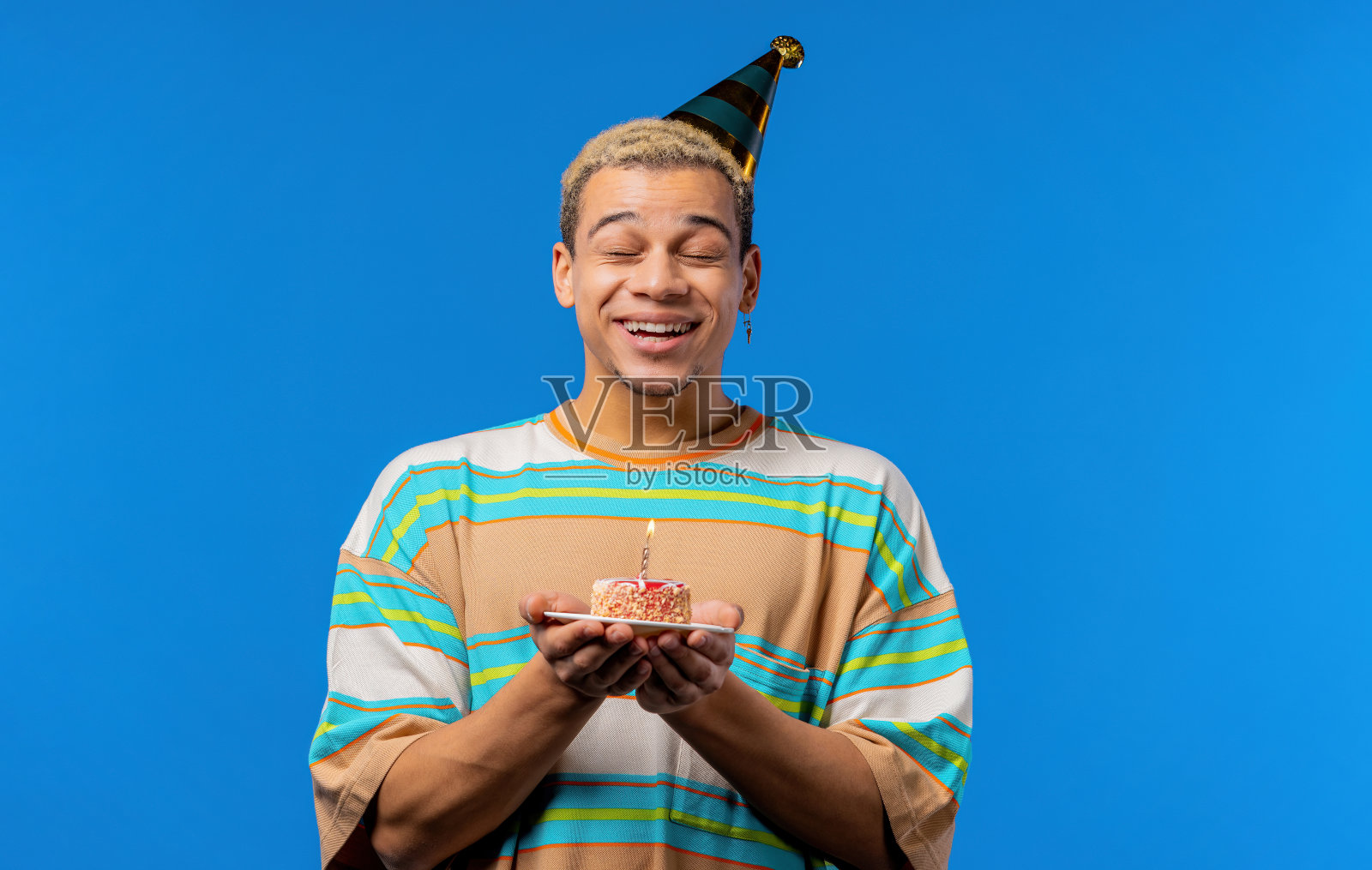 生日快乐，男人许愿-在蛋糕上吹蜡烛。卷发男笑着，庆祝纪念日。年轻的少年在蓝色背景。照片摄影图片