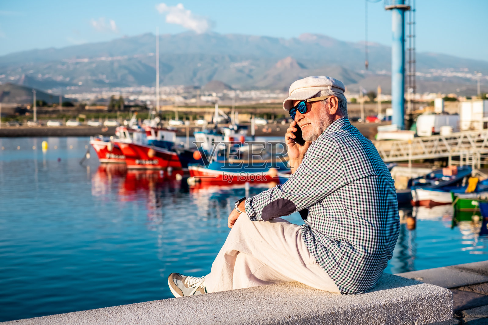 穿着休闲衬衫和帽子的英俊的高级男子坐在港口边看船和大海边打电话。放松的年长胡须男性戴着太阳镜享受旅行或退休照片摄影图片