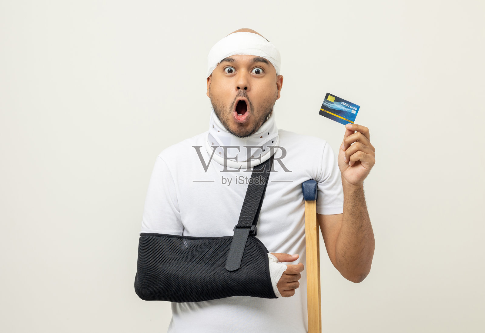 亚洲男子断了胳膊和腿，拿着信用卡付款。男子戴上石膏石膏夹板，拄着拐杖。病人佩戴带颈圈的吊带支撑臂。人身保险及意外事故照片摄影图片