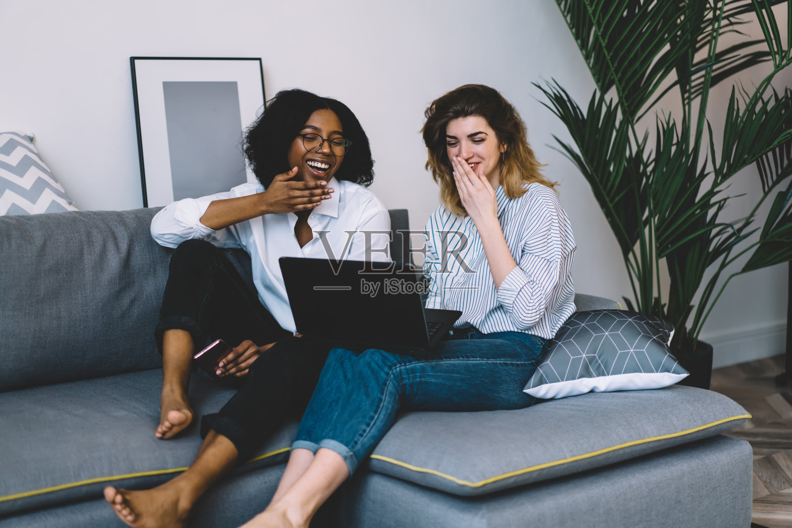 快乐的多种族同胞朋友在通过笔记本电脑应用程序浏览网页时大笑，快乐的各种时髦女孩在舒适的沙发上休息，微笑着阅读积极的上网本新闻照片摄影图片