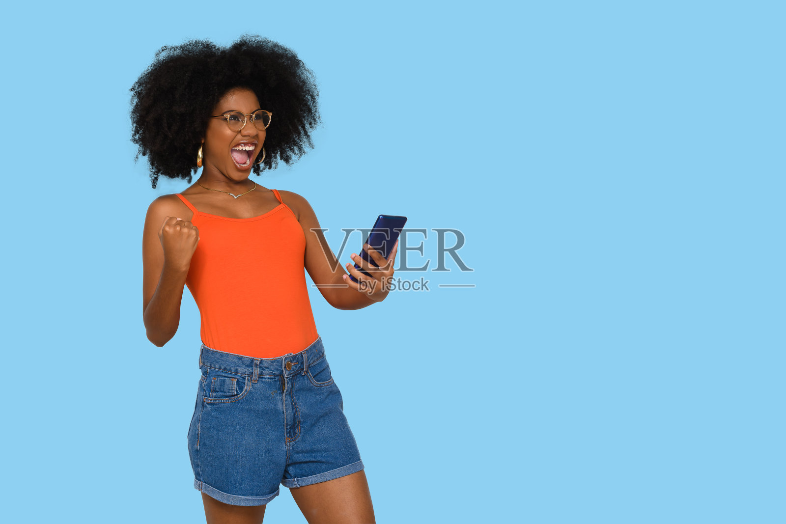 非常惊讶的年轻女子庆祝，一只手拿着智能手机，黑色的权力爆炸风格的头发在蓝色背景照片摄影图片