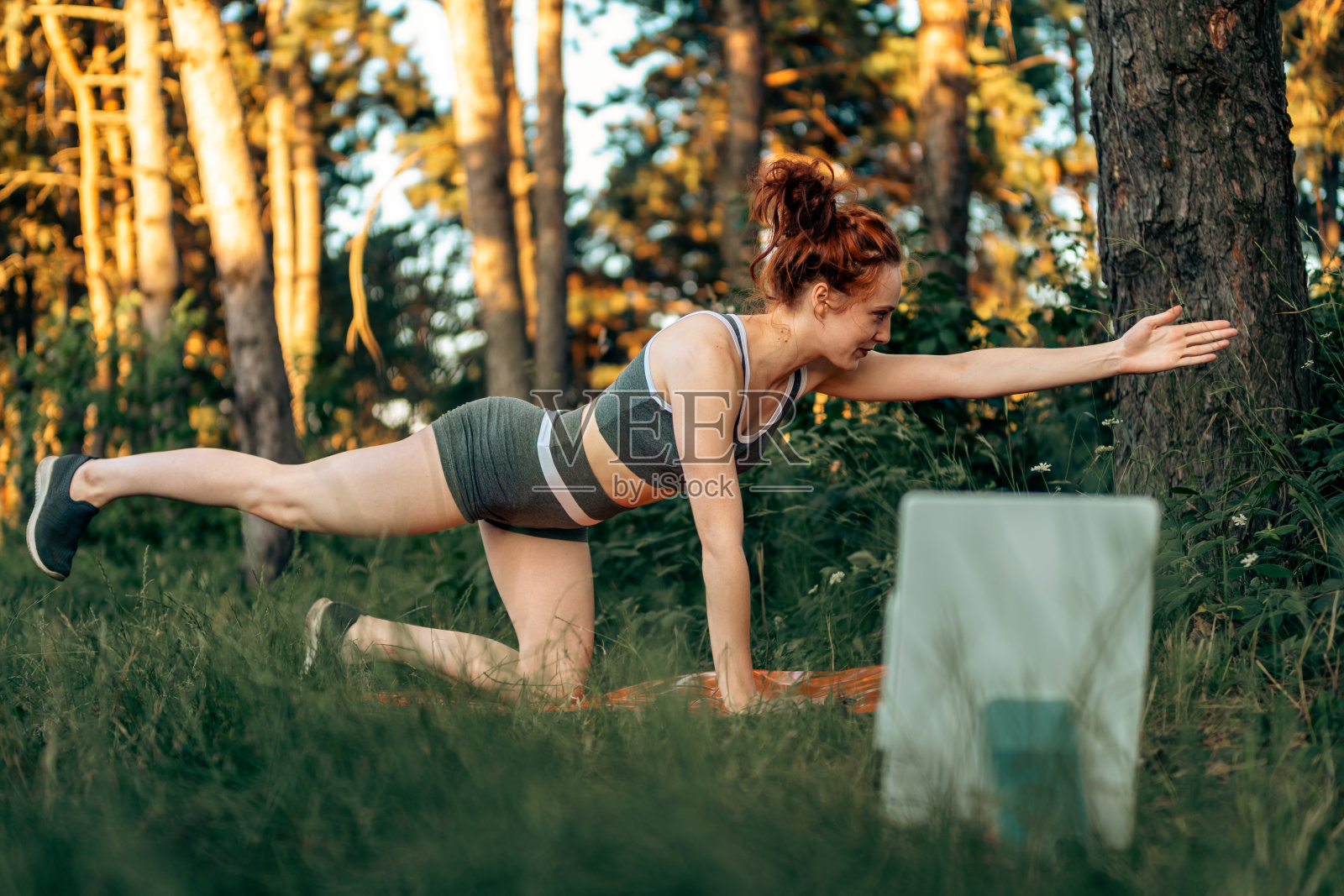 年轻的白人女子在森林里做健身拉伸瑜伽运动，同时使用平板电脑进行在线课程或虚拟教程。健康的生活方式。照片摄影图片