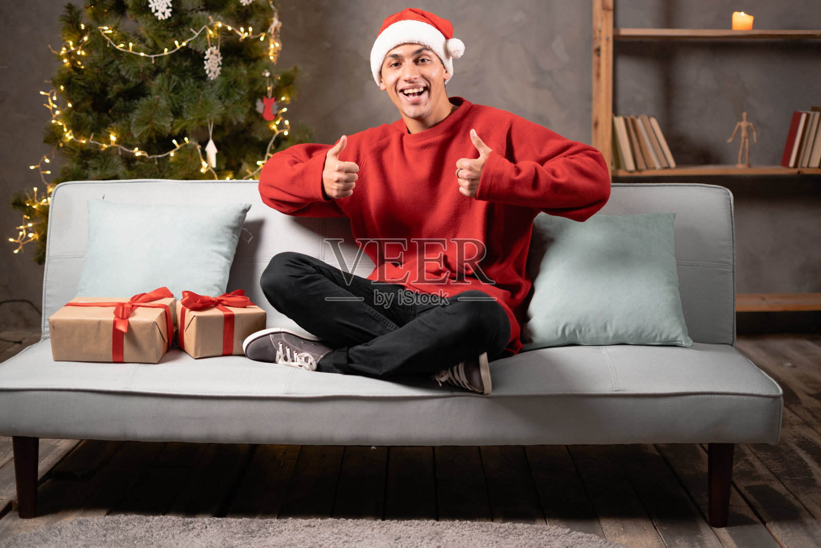 一个漂亮的有吸引力的男人的肖像在红色圣诞老人服装显示大拇指与两只手在沙发上在家里。圣诞创意概念照片摄影图片