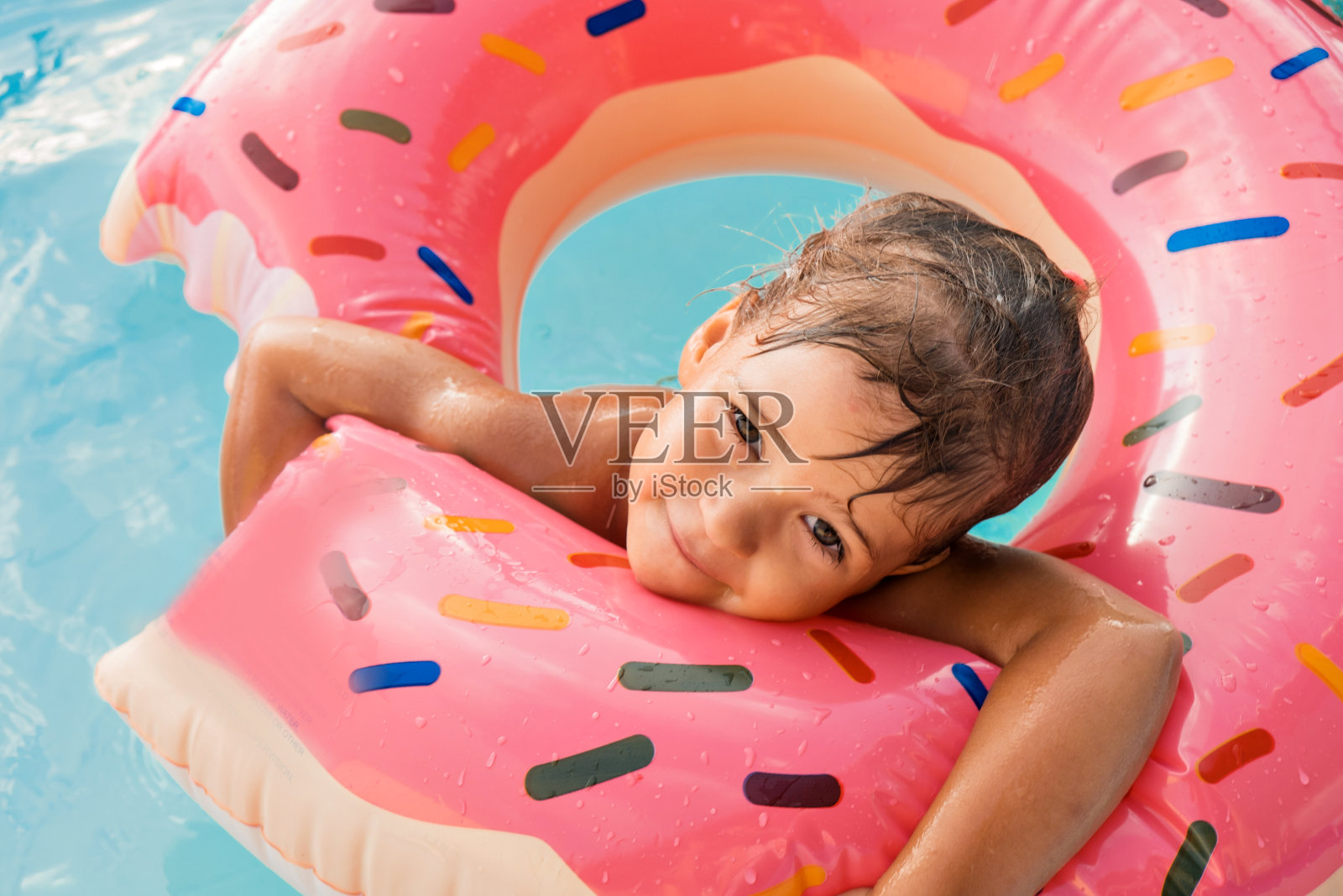 一个欧洲女孩戴着一个粉红色的大橡胶圈在蓝色的游泳池里洗澡。湿漉漉的头发和幸福的微笑。海浪、酒店和海滩。一家人正在享受一个快乐的假期。照片摄影图片