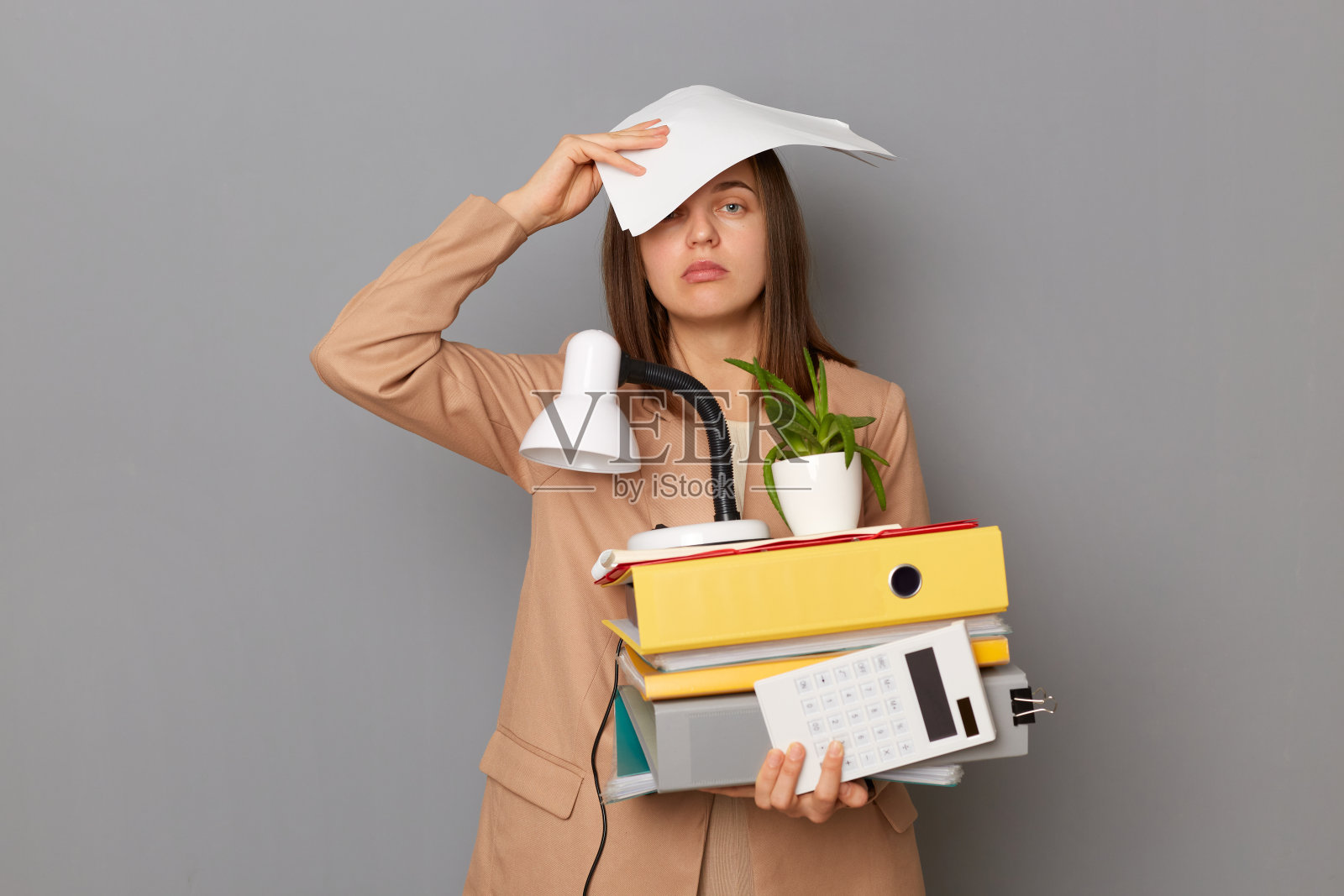 室内镜头，疲惫疲惫的女人穿着时尚的米色夹克，拿着办公室的东西，在灰色的背景上摆出孤立的姿势，闭着眼睛站着，她的工作有问题。照片摄影图片