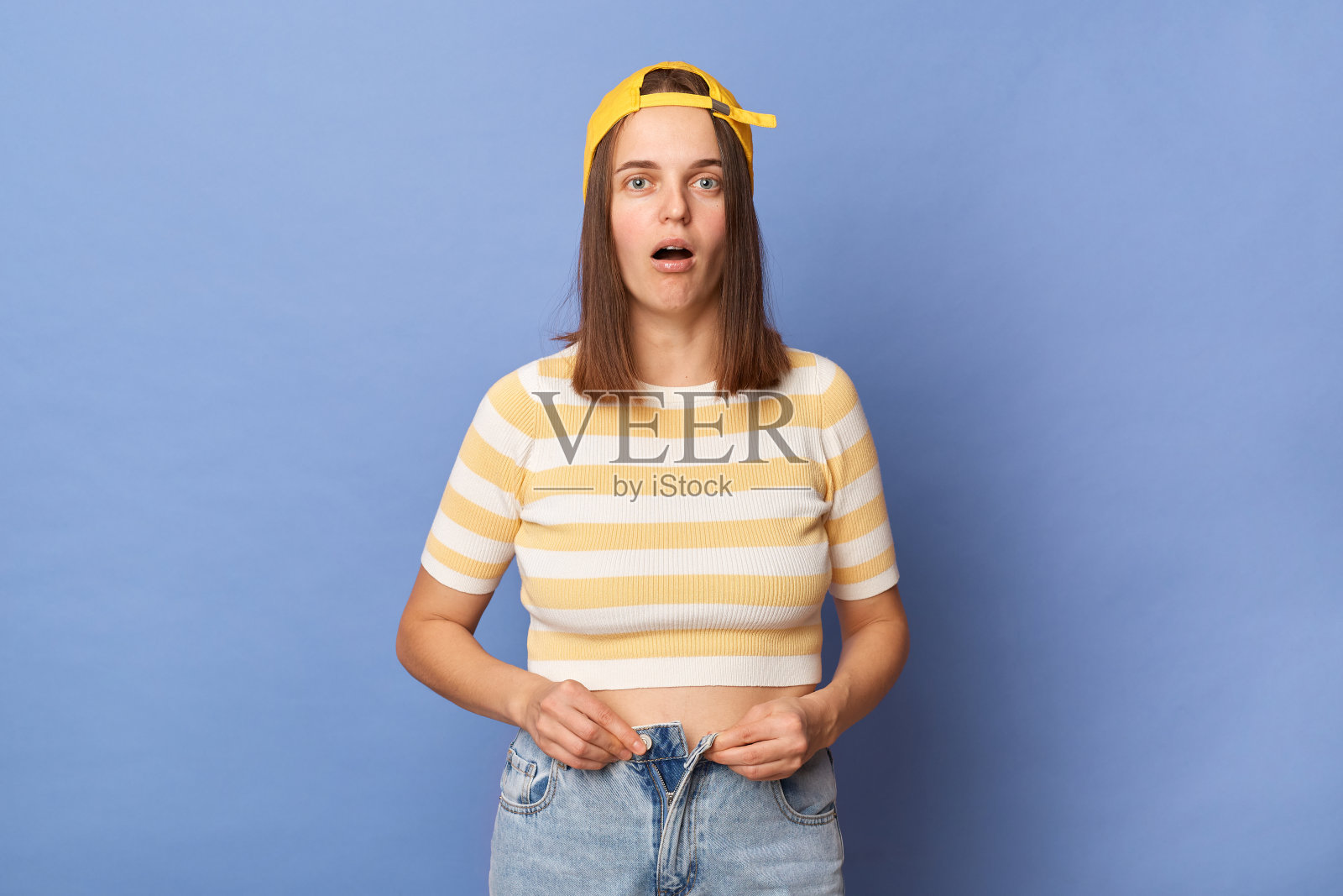 震惊悲伤的少女穿着条纹t恤和棒球帽站在蓝色背景孤立的肖像，女性看着相机，需要减肥。照片摄影图片
