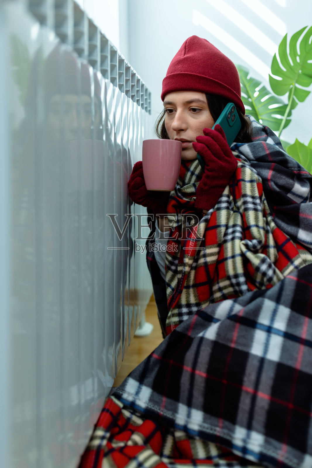一名年轻女子因公寓内的气温过低而感到不快，她一边打电话给一个充满活力的服务公司，一边在暖气片旁喝茶。照片摄影图片