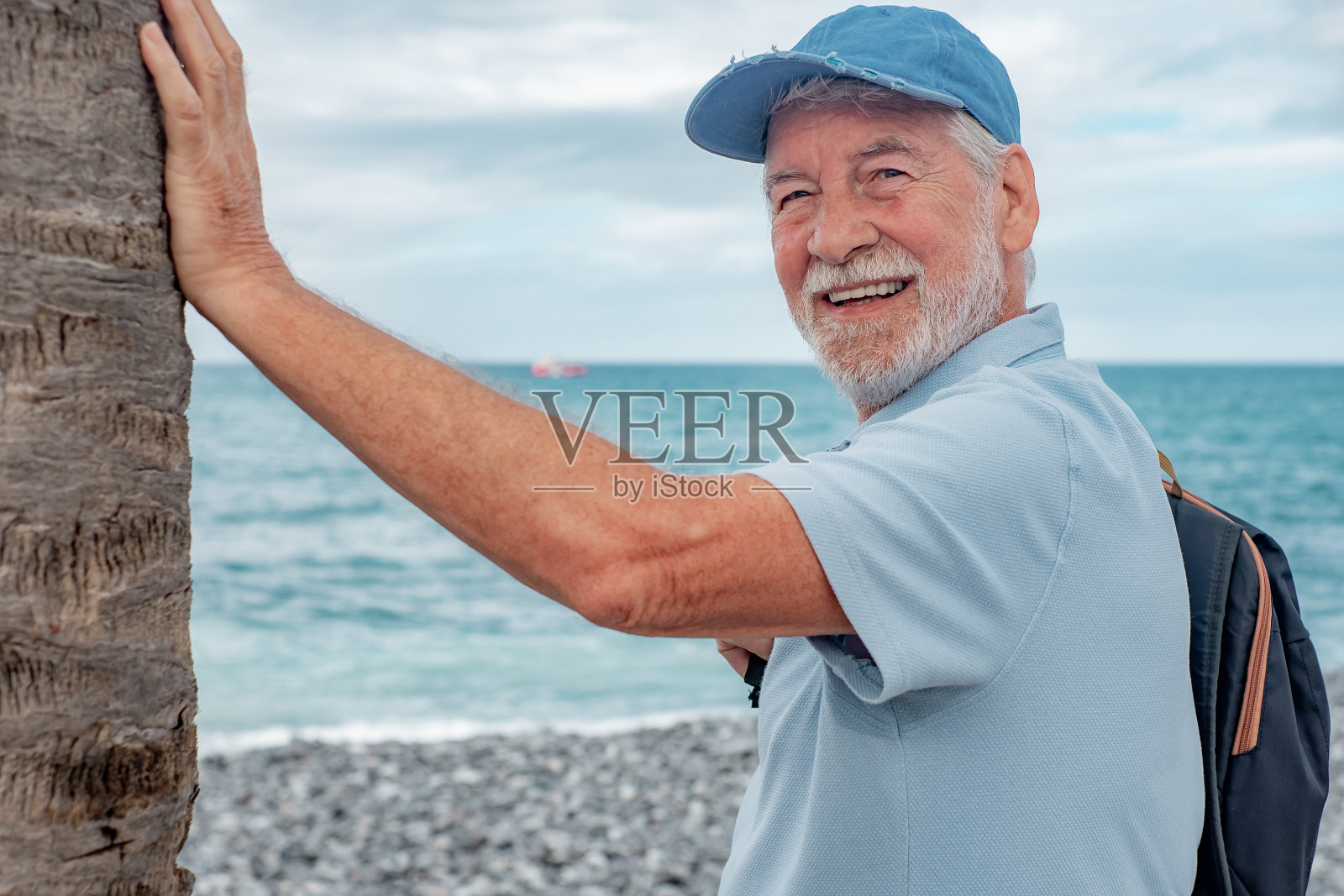 微笑的高级男子站在海滩上，靠近一棵棕榈树，享受空闲时间假期或退休。有吸引力的胡子男子与帽子和白发看着相机。水面上的地平线照片摄影图片