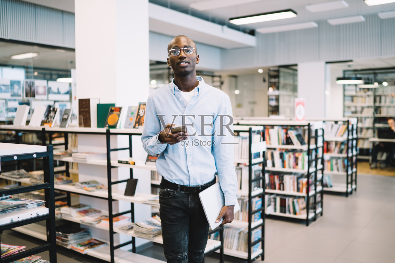 微笑的黑人男子靠近书架，用智能手机和书看着相机照片摄影图片