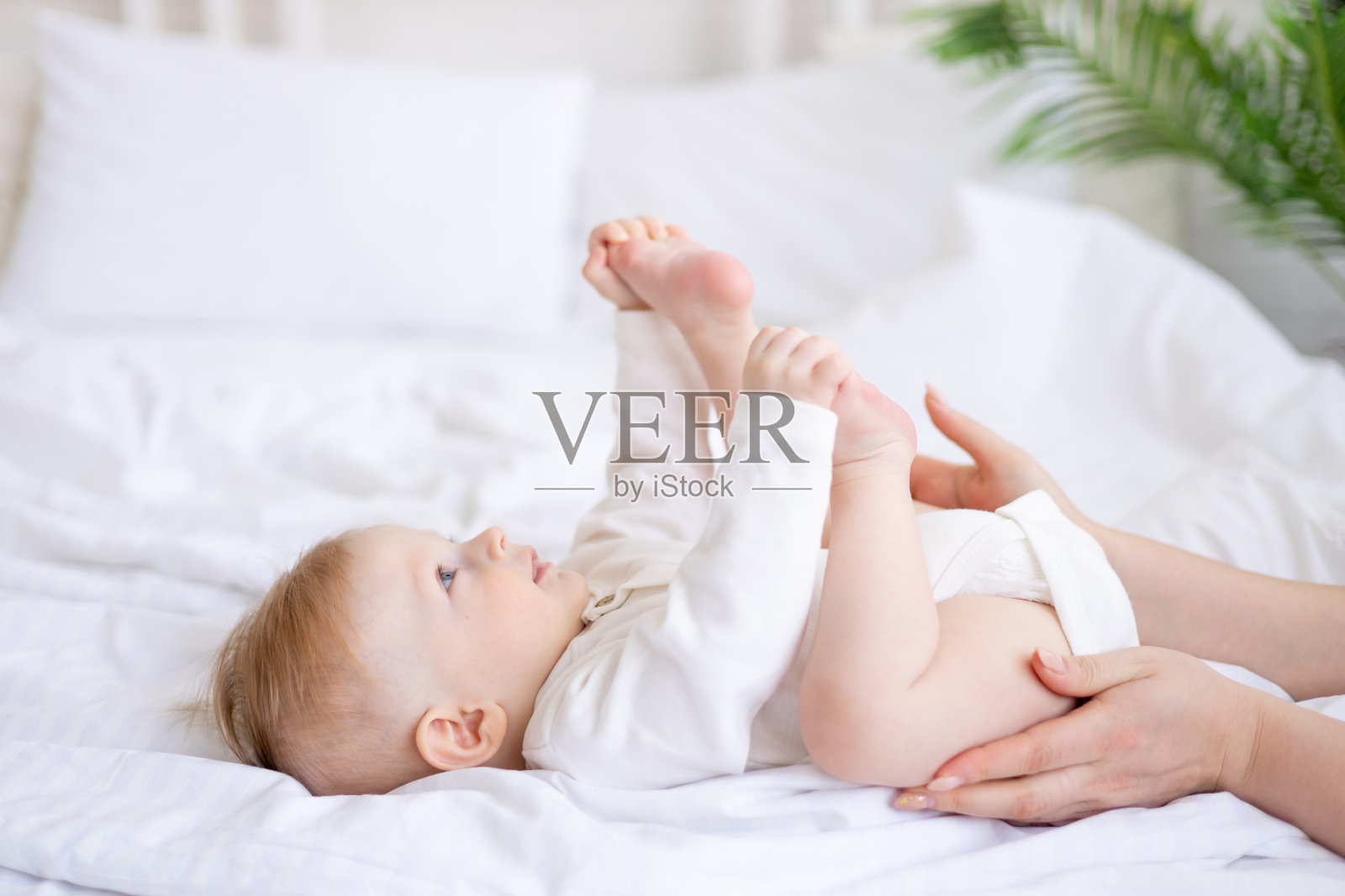 妈妈的手牵着宝宝的腿，6个月大的金发男孩在明亮的卧室里睡在白色的床上，早上穿着棉质的紧身衣，概念儿童用品照片摄影图片