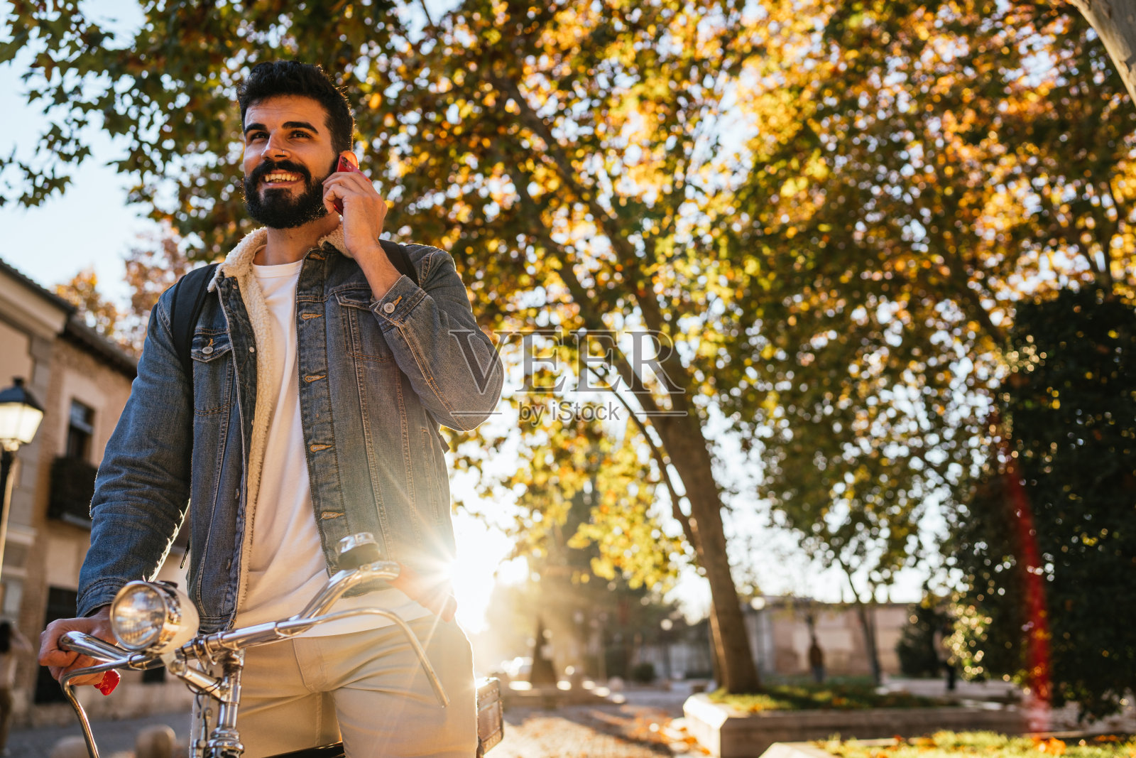 日落时分，年轻男子穿着牛仔夹克和背包，骑在他的经典复古自行车上，一边打电话。照片摄影图片