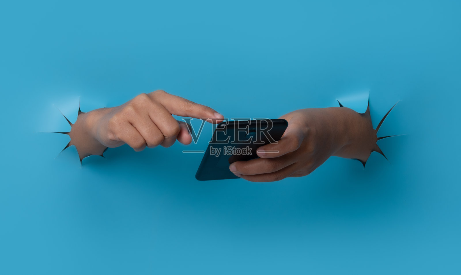 女性手握智能手机，触屏突破蓝色纸背景。照片摄影图片