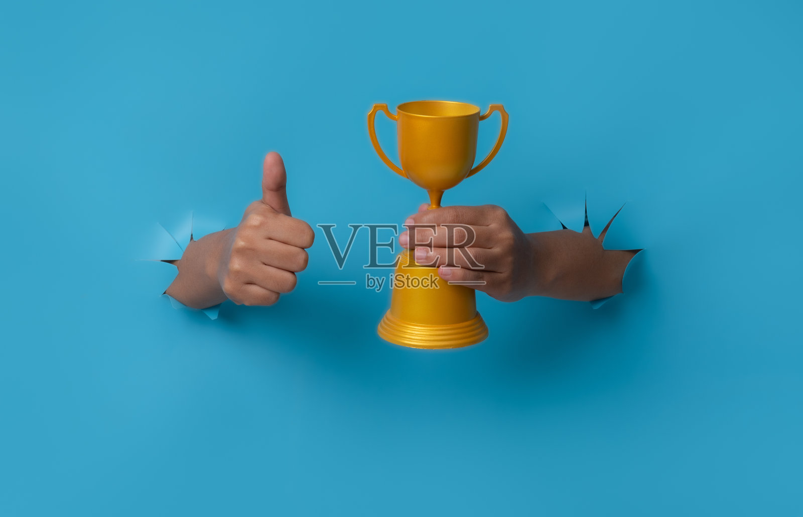 手持冠军金杯的女性手从蓝色的纸背景中突破。照片摄影图片