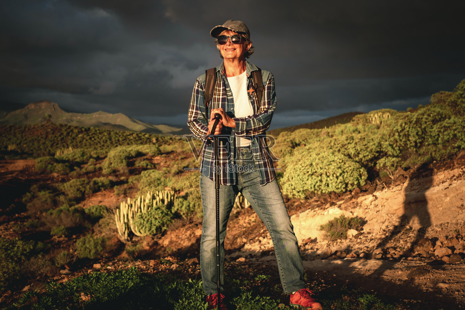 美丽的高级妇女在帽子和运动服走在大自然夕阳下享受自由和假期。快乐的成熟女性用棍子看着山地热带景观照片摄影图片