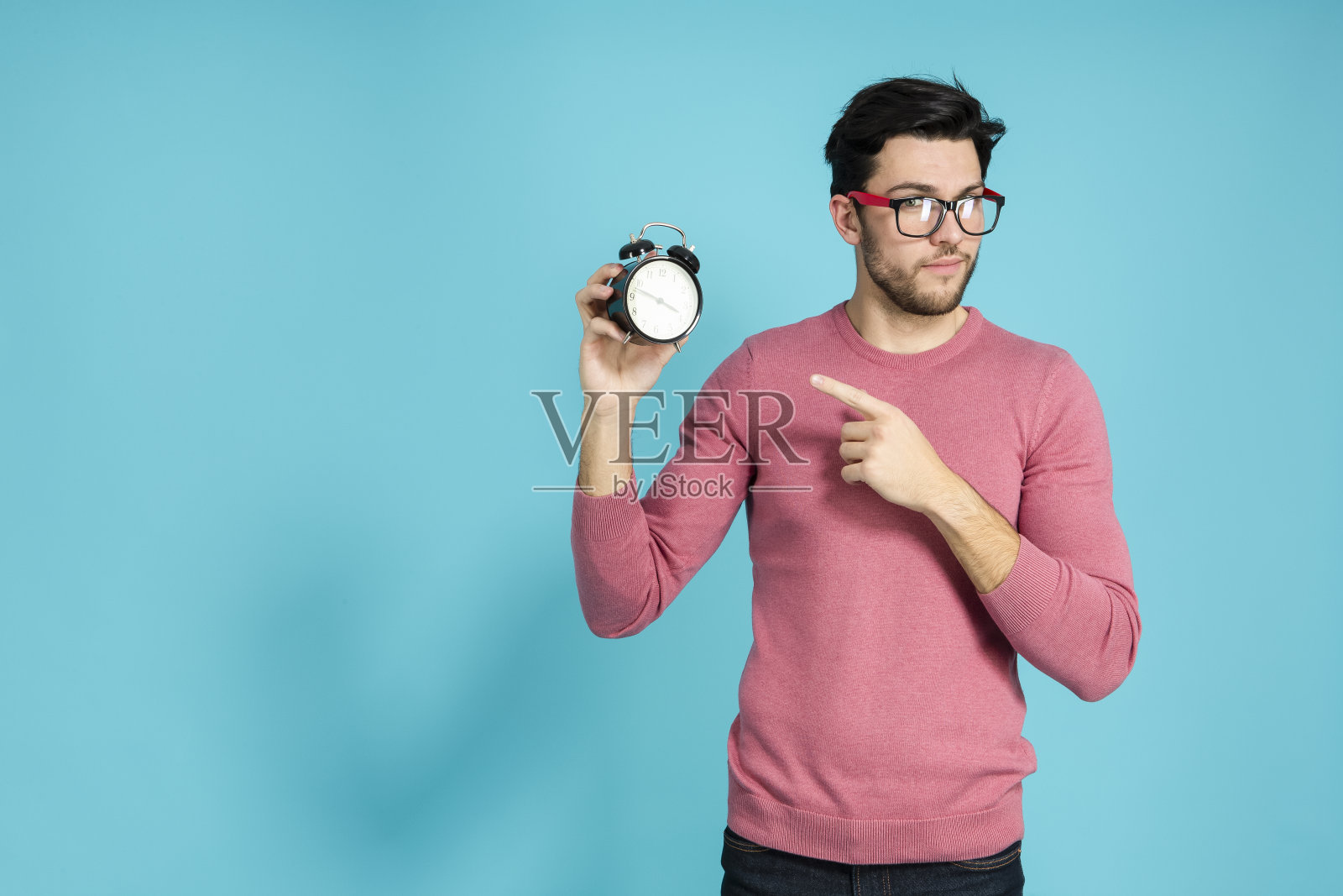 年轻的白人男子在眼镜的自然肖像手势与闹钟和举起手与尖尖的手指。照片摄影图片