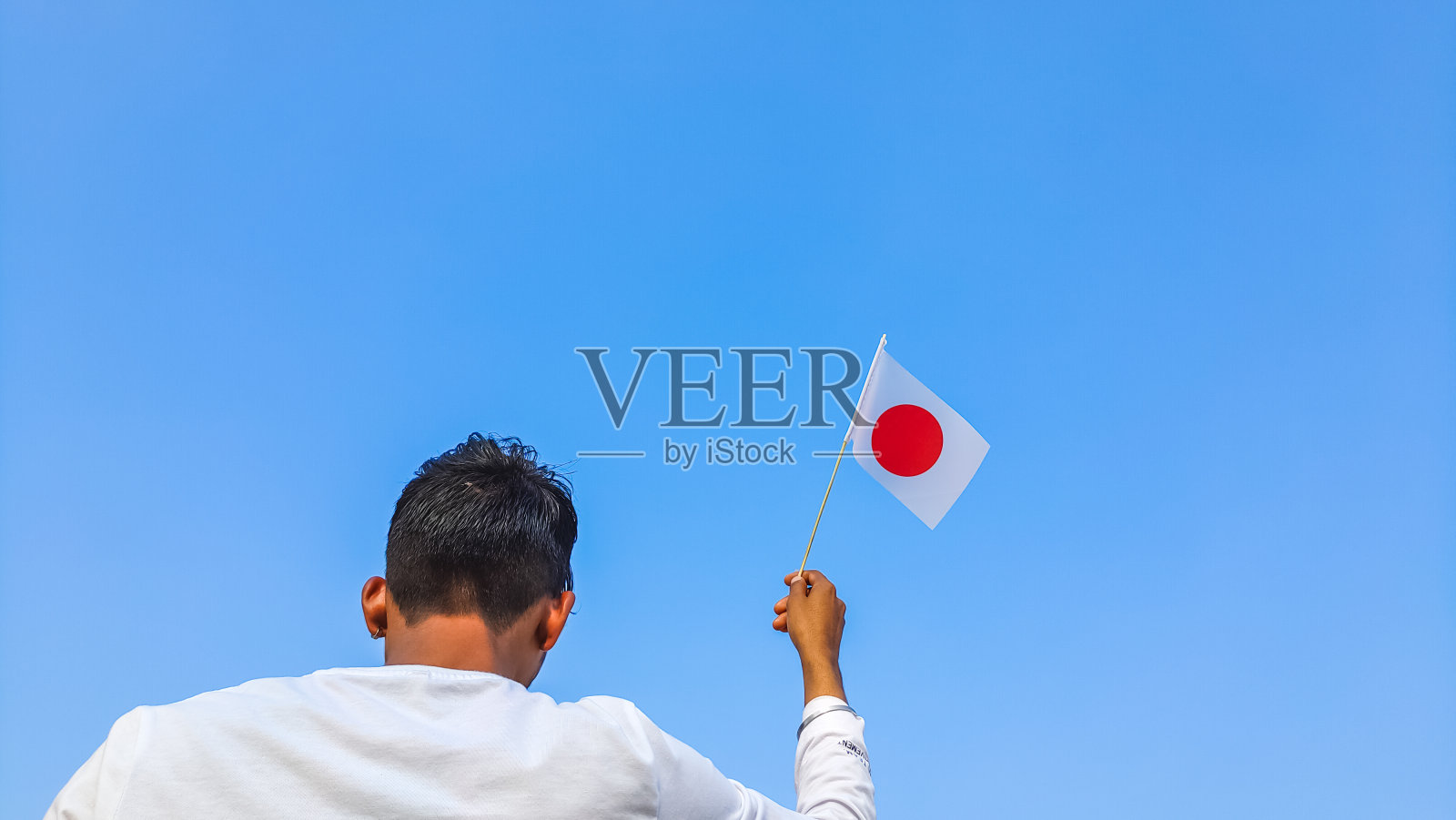 男孩在湛蓝的天空下举着日本国旗。男子手挥舞日本国旗从后面看，复制空间照片摄影图片